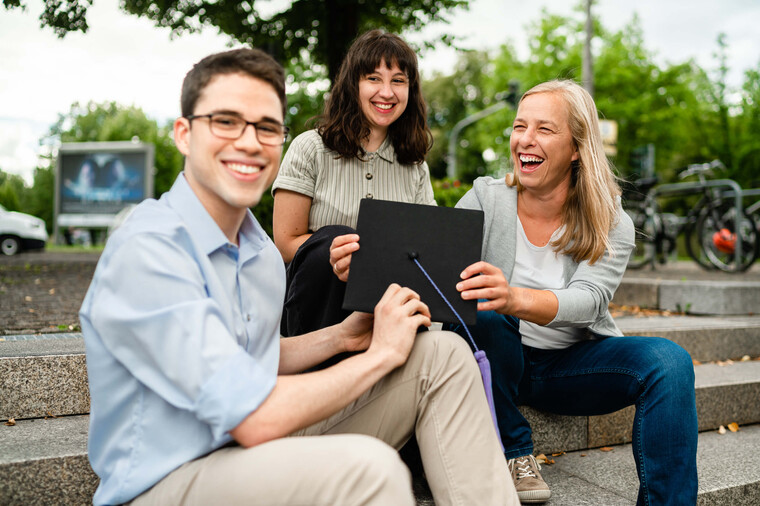 Drei junge Menschen sitzen lachend im Freien auf dem Campus und schauen auf ein Tablet.